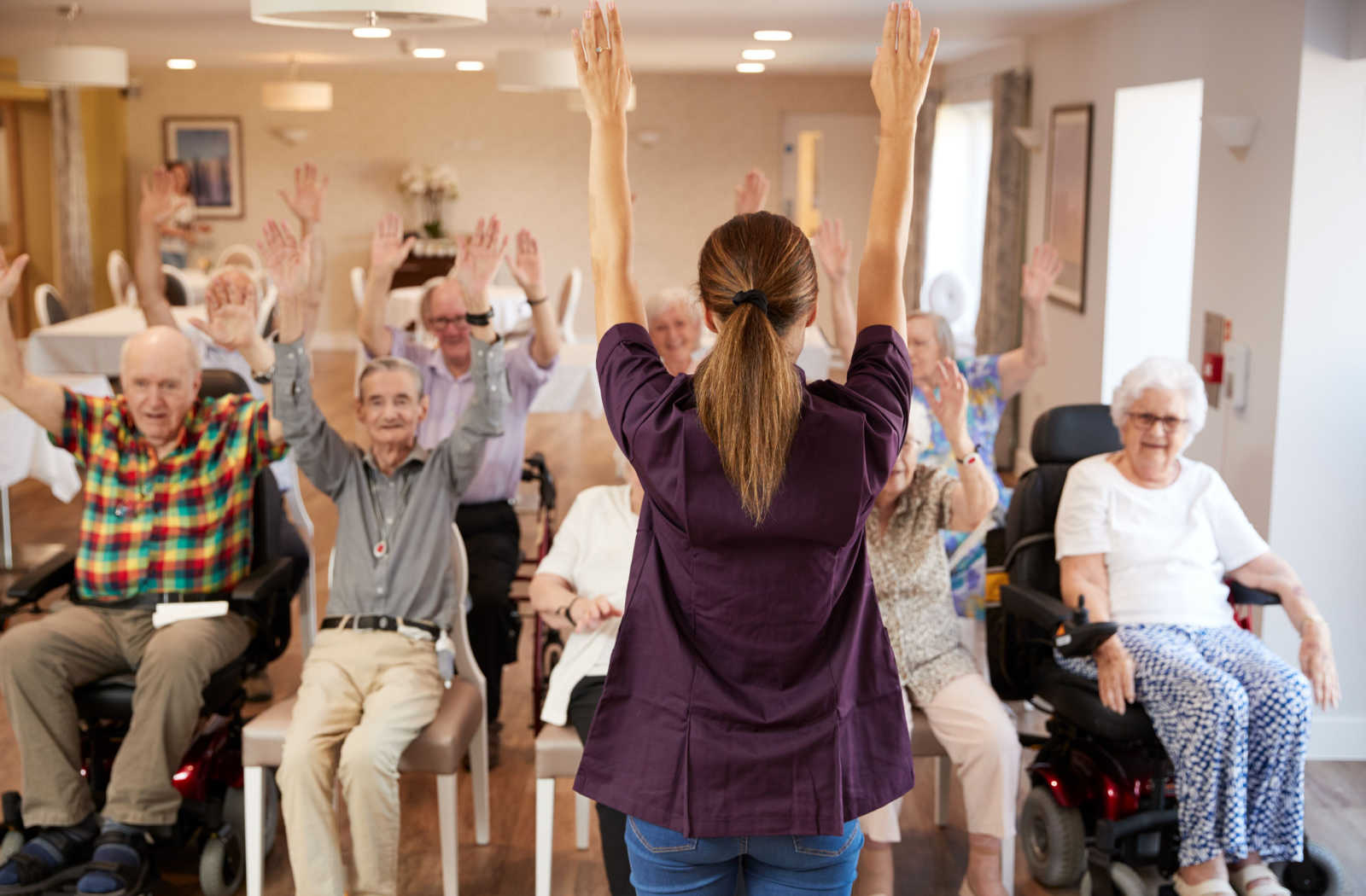 A group of seniors in a senior living community sitting and exercising together, guided by a trained staff member.
