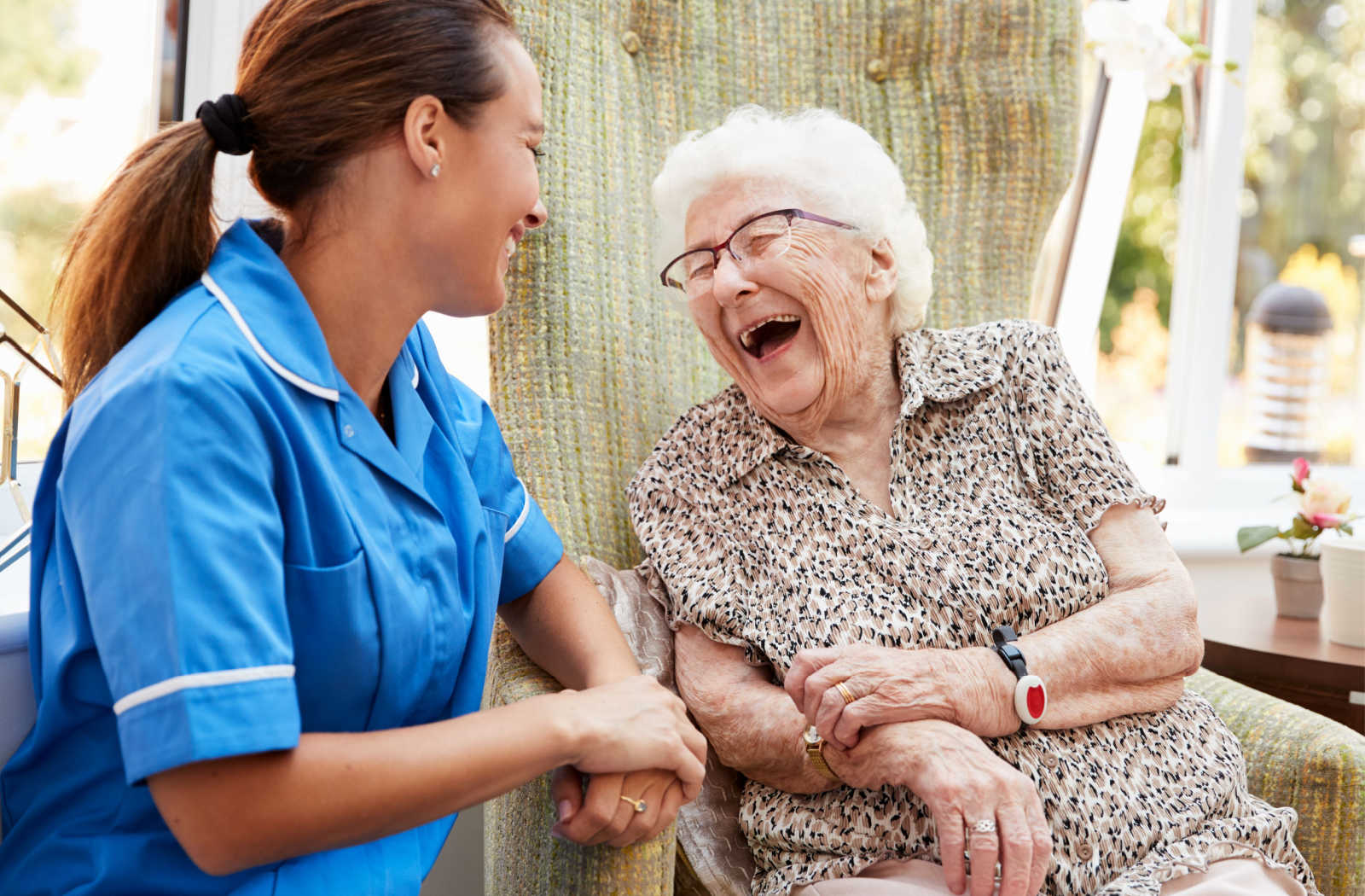 A senior woman in a senior living community sitting on a chair smiling and laughing with a young female nurse.