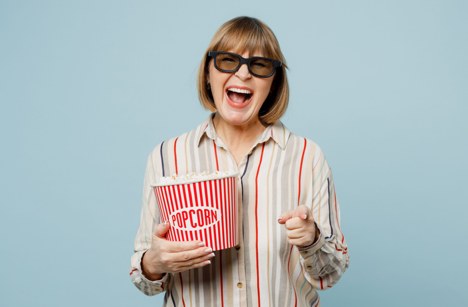 Senior woman in a striped shirt holding a bucket of popcorn laughing and pointing towards the camera.